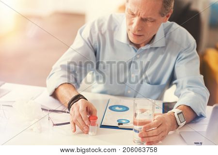 It wil help. Cheerless professionla mature male office worker sitting at the table and going to take medicines while sitting in the office