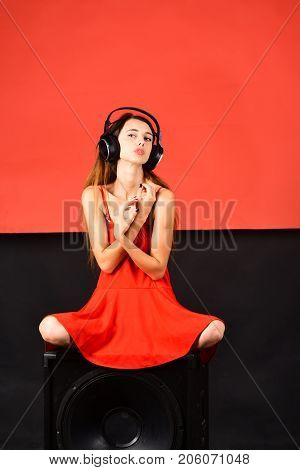 Dj sits on boombox on black and red background. Girl with loose hair wears headphones and red dress. Pleasure music and creative lifestyle concept. Technologies and music concept.