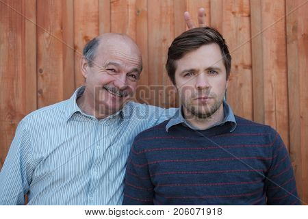 Father pranking his son with bunny ears isolated on wooden background. Young nman is very serious, old gentleman smiles.