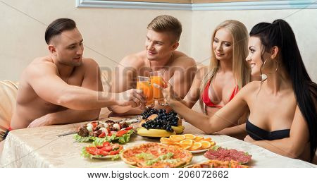 Shirtless muscular men and their girl friends in bicinis sitting at table with fruit, shashlik and drinks in sauna talking and laughing together