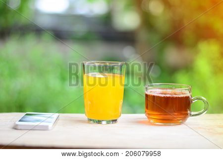 A Glass Of Orange Juice ,a Cup Of Tea And White Mobliephone On Wooden Table In Garden