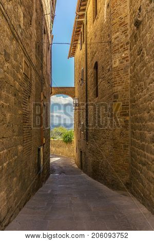 Vertical photo with view into small narrow street. The buildings are build from volcanic tuf stone typical for Tuscany in Italy. The view between houses shows the countryside with blue sky.