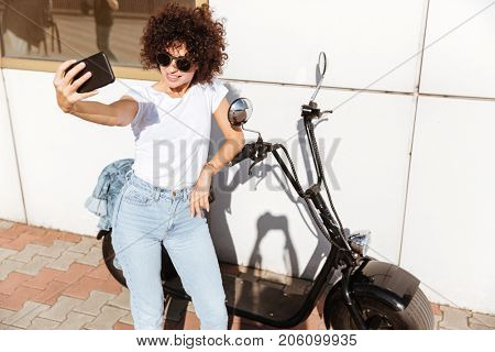Stylish pretty girl in sunglasses taking a selfie while leaning on a motorbike outdoors