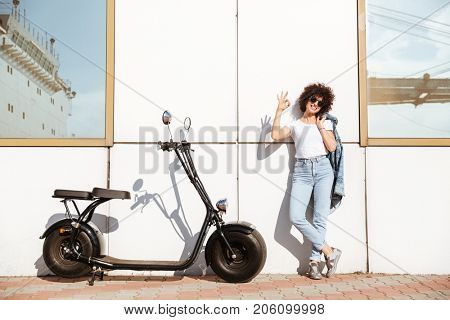 Attractive stylish woman in sunglasses posing while standing against a wall outdoors with a motorbike and showing ok gesture