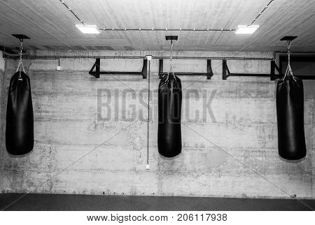 Three black punching bags in the empty boxing gym with naked grunge wall in background black and white