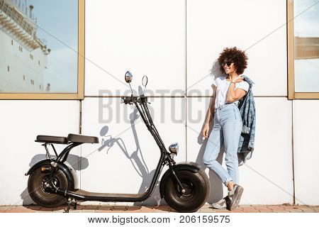 Young attractive woman in sunglasses posing while standing against a wall outdoors with a motorbike