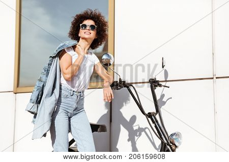 Pretty young woman in sunglasses posing while standing against a wall outdoors with a motorbike