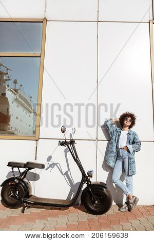 Young pretty woman in sunglasses posing while standing against a wall outdoors with a motorbike