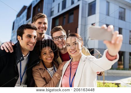 business, education and corporate concept - international group of people with conference badges taking selfie by smartphone on city street