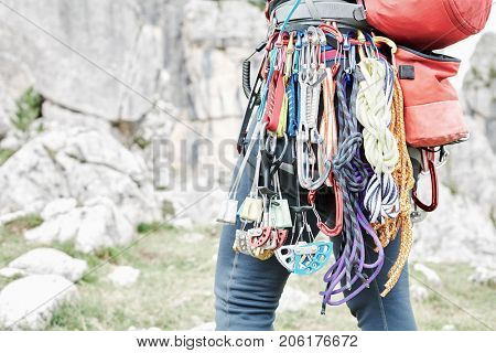 Close-up of mountaineer with trad climbing rack including backpack, chalk bag, harness with spring-loaded cams, nuts, quickdraws, slings and carabiners preparing for ascent in summer mountains