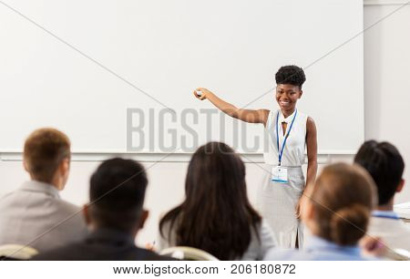 business, education and people concept - smiling african american businesswoman or teacher with remote and group of students at conference presentation or lecture