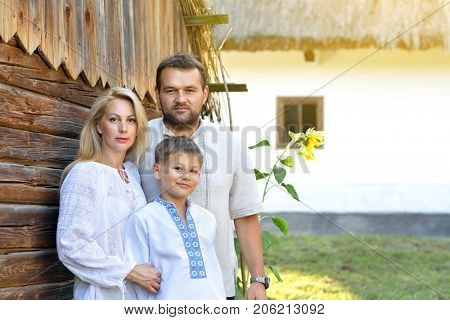 Ukrainian family in traditional clothing vyshyvanka over landscape of tipical ucrainian village, summer outdoor. Ukrainian people posing near the house. Slavic mother, father and son