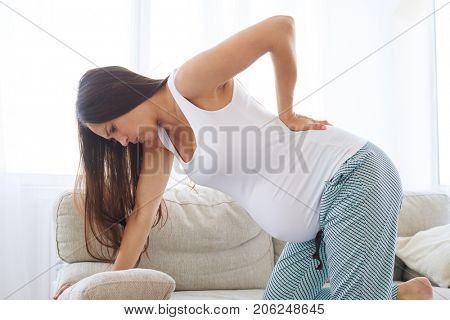 Side view of young pregnant woman having backpain, leaning on sofa 