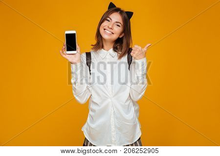 Smiling teenage schoolgirl in uniform with backpack holding blank screen mobile phone and showing thumbs up gesture isolated over orange background