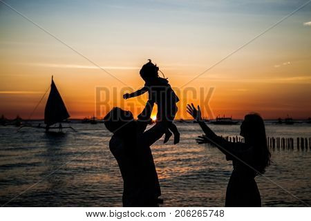 Happy family - silhouette of father tossing baby girl and mother on sunset sea . Philiphines.