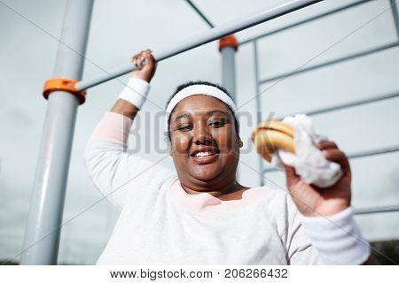 Happy young obese female with hamburger holding by bar of outdoor sport facilities and going to eat