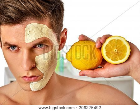 Facial mask from fresh fruits and clay for man concept. Face with treatment mud applied. Male holding lemon half for skin care procedure in salon. Removing black dots.