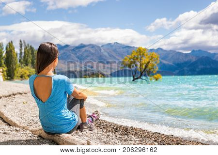 New Zealand travel tourist woman looking at Wanaka lone tree lake landscape. Girl relaxing enjoying view sitting on beach at Wanaka nature mountains outdoors.