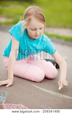 Sunny and warm summer days and happy chilhood lisure time. Cute little girl drawing with chalk crayons at the asphalt alone. Summer vacation.