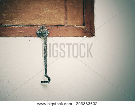 Old metal shutter hook on edge of brown wooden window; unfastened and hanging vertically in front of white wall. Vintage and cinematic look with vignette. Right copy space for add text.