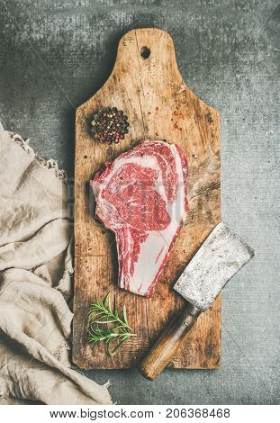 Flat-lay of raw prime beef meat dry-aged steak rib-eye on bone with seasoning and chopper knife on wooden cutting board over grey concrete background, top view. Meat high-protein dinner concept