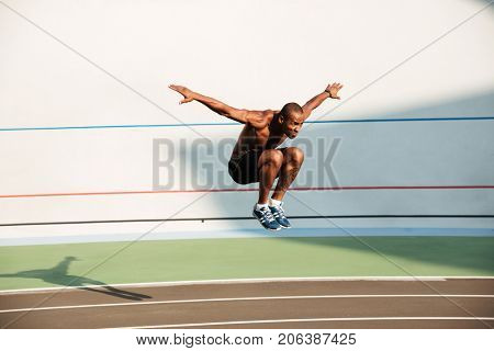 Full length portrait of a half naked strong fit african sportsman jumping at the track field outdoors