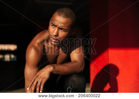 Close up portrait of a muscular half naked african sportsman sitting on his knee and looking away indoors