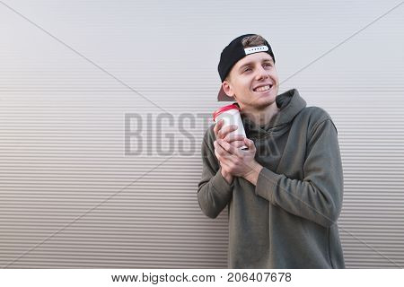 A sweet and positive young man with coffee in his hands hugs a cup against a background of a light wall.