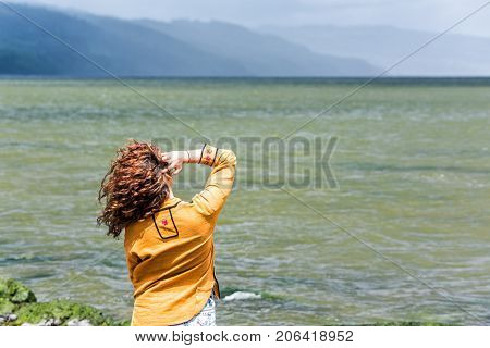 Back Of Young Woman Looking At Stormy Dark Green Ocean Or River Fixing, Touching Curly Hair Blowing