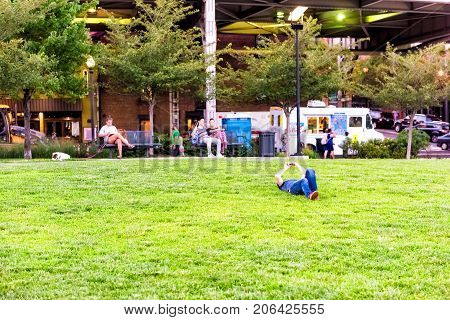 Washington DC USA - August 4 2017: People sitting on benches in green downtown Georgetown waterfront park neighborhood with young man lying on grass lawn in evening with smartphone phone