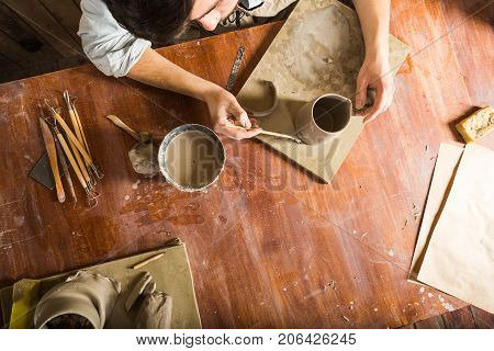 pottery, stoneware, ceramics art concept - workplace of potter, craftsman bent over clay cup with tools, male hands connecting pieces of fireclay, young male sits at a workshop behind table, flat lay