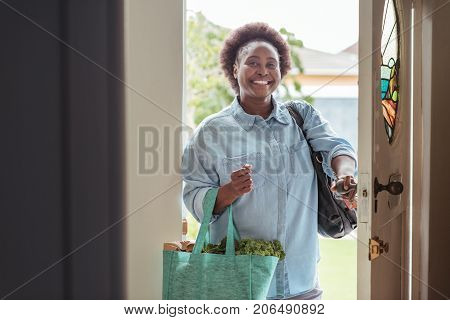 Smiling African woman walking through the front door of her home with a shopping bag full of fresh produce