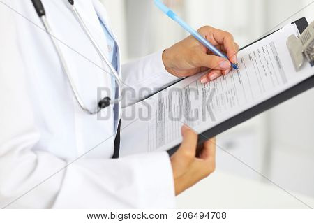 Close-up of a female doctor filling  up medical form at clipboard while standing straight in hospital.