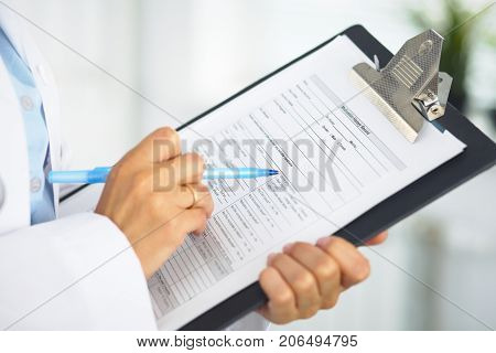 Close-up of a female doctor filling  up medical form at clipboard while standing straight in hospital.