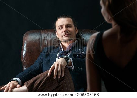 The actor or mature man wearing suit in a classical retro fashion style. The stylish man sitting on a leather couch in a studio. The woman standing near