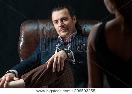 The actor or mature man wearing suit in a classical retro fashion style. The stylish man sitting on a leather couch in a studio. The woman standing near