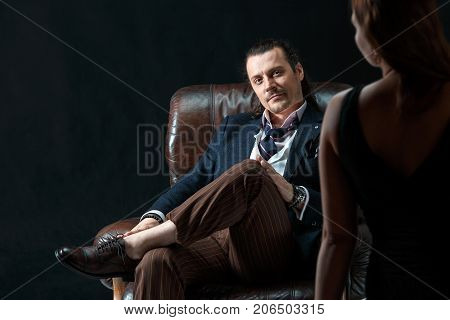 The actor or mature man wearing suit in a classical retro fashion style. The stylish man sitting on a leather couch in a studio. The woman standing near