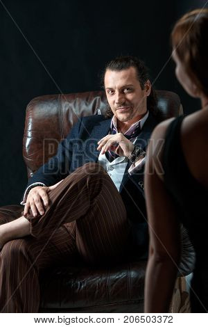 The actor or mature man wearing suit in a classical retro fashion style. The stylish man sitting on a leather couch in a studio. The woman standing near