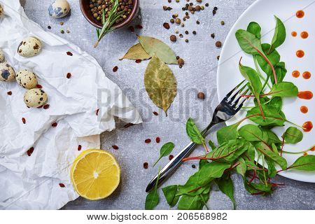 Top view of a white plate with drops of sauce and fresh green leaves, bay leaves, a half of juicy yellow lemon, little speckled quail eggs, a jar with different seasonings on a light gray background.