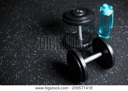 A blue plastic bottle for water, two heavy metal dumbbells, equipment for sports competitions, gyms, trainings, routines on a dark blurred background.