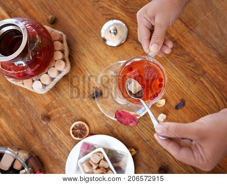 Top view of a bowl of crunchy biscuits with nuts and spices on a wooden background. Woman's hands holding a spoon with brown sugar and a cup of red, hot strawberry tea. A big and full tea kettle.
