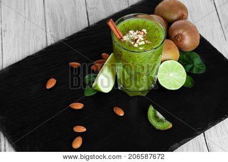 A thick, blended kiwi milkshake with juicy limes and cut apples on a black tray. A fresh non-alcoholic beverage with spices and nuts on the wooden table background. Pretty natural composition.