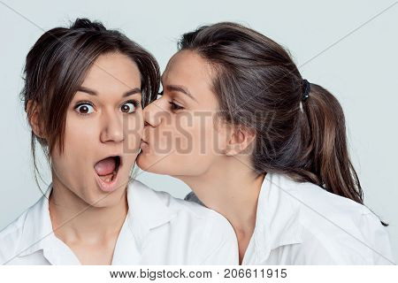 Studio portrait of young female twins sisters on gray. The one girl kissing another on the cheek