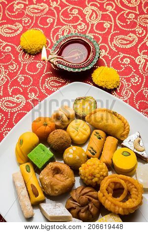 stock photo of Indian sweet or mithai and oil lamp or diya with gift box and flowers on decorative or colourful background, selective focus