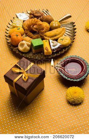 stock photo of Indian sweet or mithai and oil lamp or diya with gift box and flowers on decorative or colourful background, selective focus