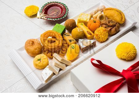 stock photo of Indian sweet or mithai and oil lamp or diya with gift box and flowers on decorative or colourful background, selective focus