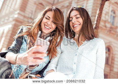Two young beautiful women friends in town using phone listening music and have a fun.