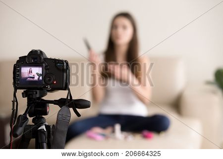 Young female videoblogger sitting on sofa, records commercial beauty product review for personal video blog. Vlogger uses contemporary photo camera to create footage for popular internet vlog at home.
