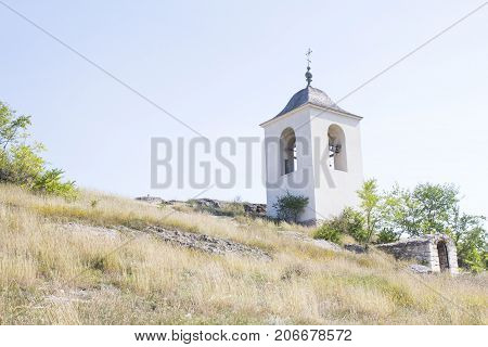 A temple on the slope near green tree and golden grass in sunny day. Orthodox church with cross on the bell tower. View from the bottom up.  Concept of credo and religion.