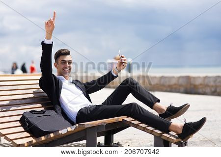 Energetic businessman enjoying work on trip and sitting on comfortable wooden bench. Emotional successful young man using smartphone and looking at camera. Winning concept